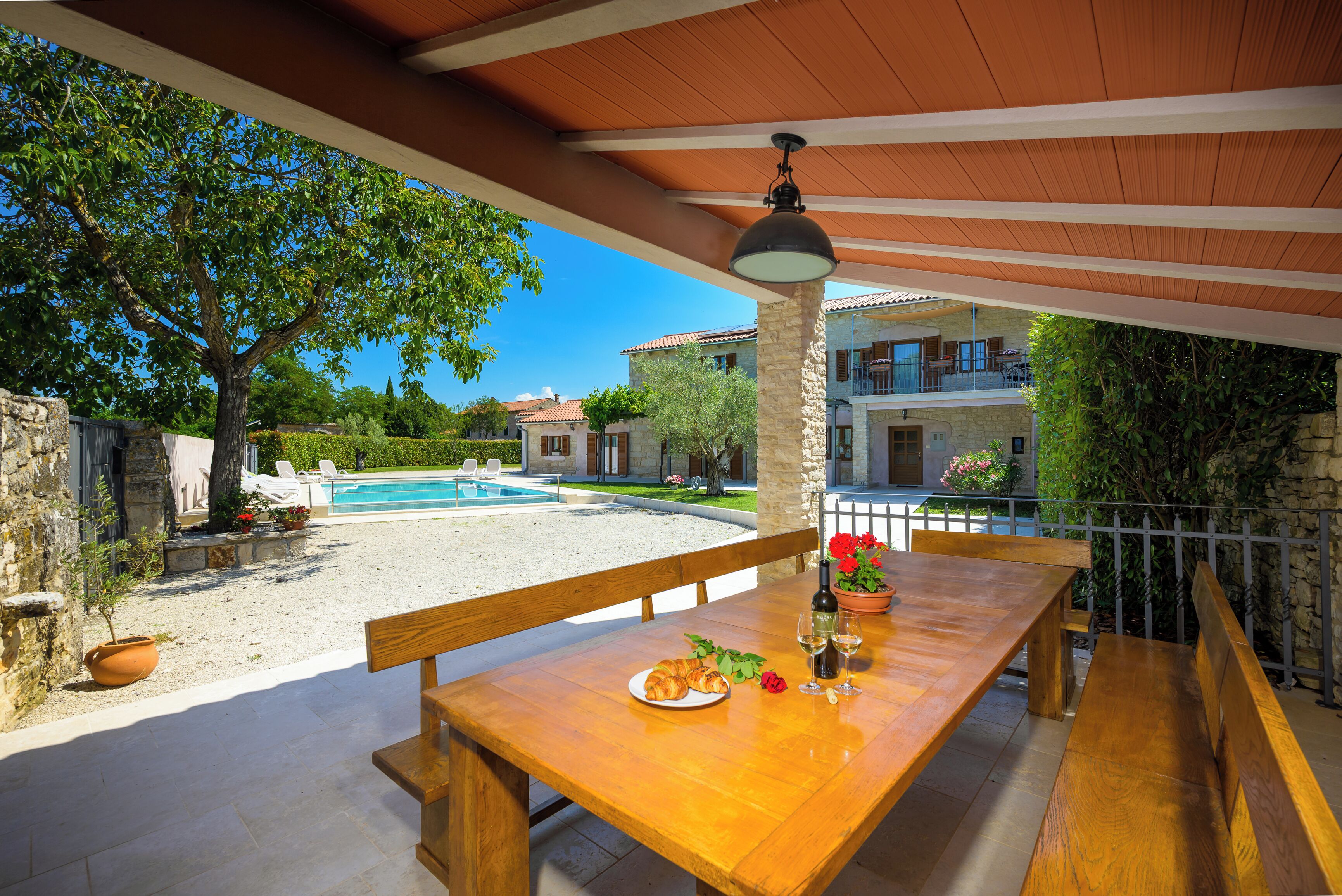 Un tavolo da pranzo in legno all'aperto con una bottiglia di vino, bicchieri e un piatto di croissant, con sullo sfondo una piscina e un edificio in pietra.