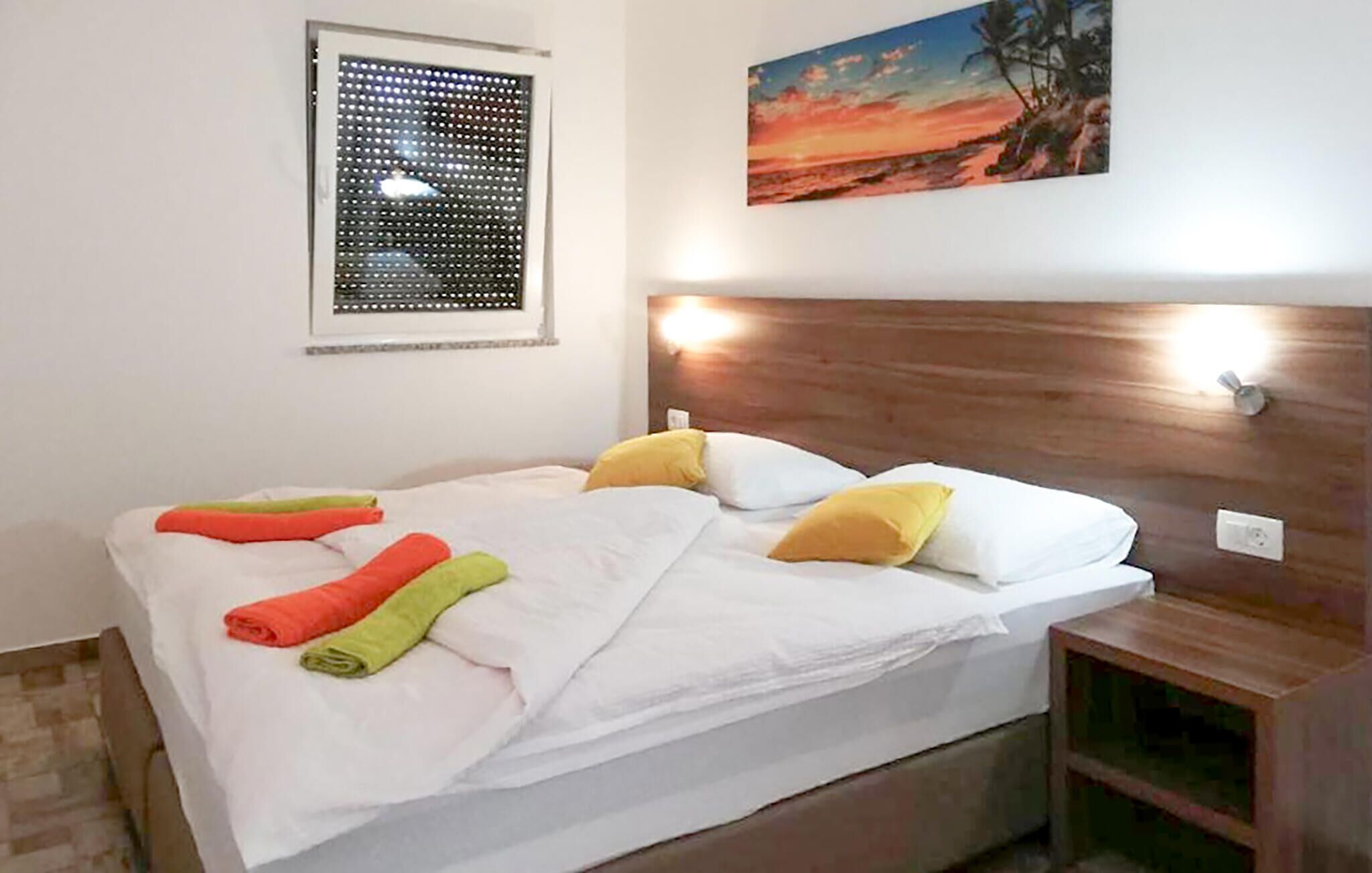 A neatly made bed with white linens and colorful towels, a wooden headboard with wall-mounted lights, a window with blinds, and a painting of a sunset.