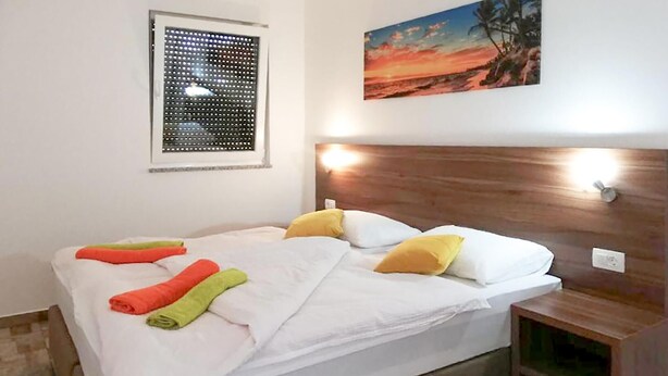 Una cama bien hecha con sábanas blancas y toallas coloridas, un cabecero de madera con luces fijadas a la pared, una ventana con persianas y un cuadro de un atardecer.