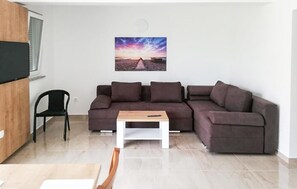 Ein Wohnzimmer mit einem Ecksofa, einem kleinen Tisch, einem Sessel und einem an der Wand montierten Fernseher.