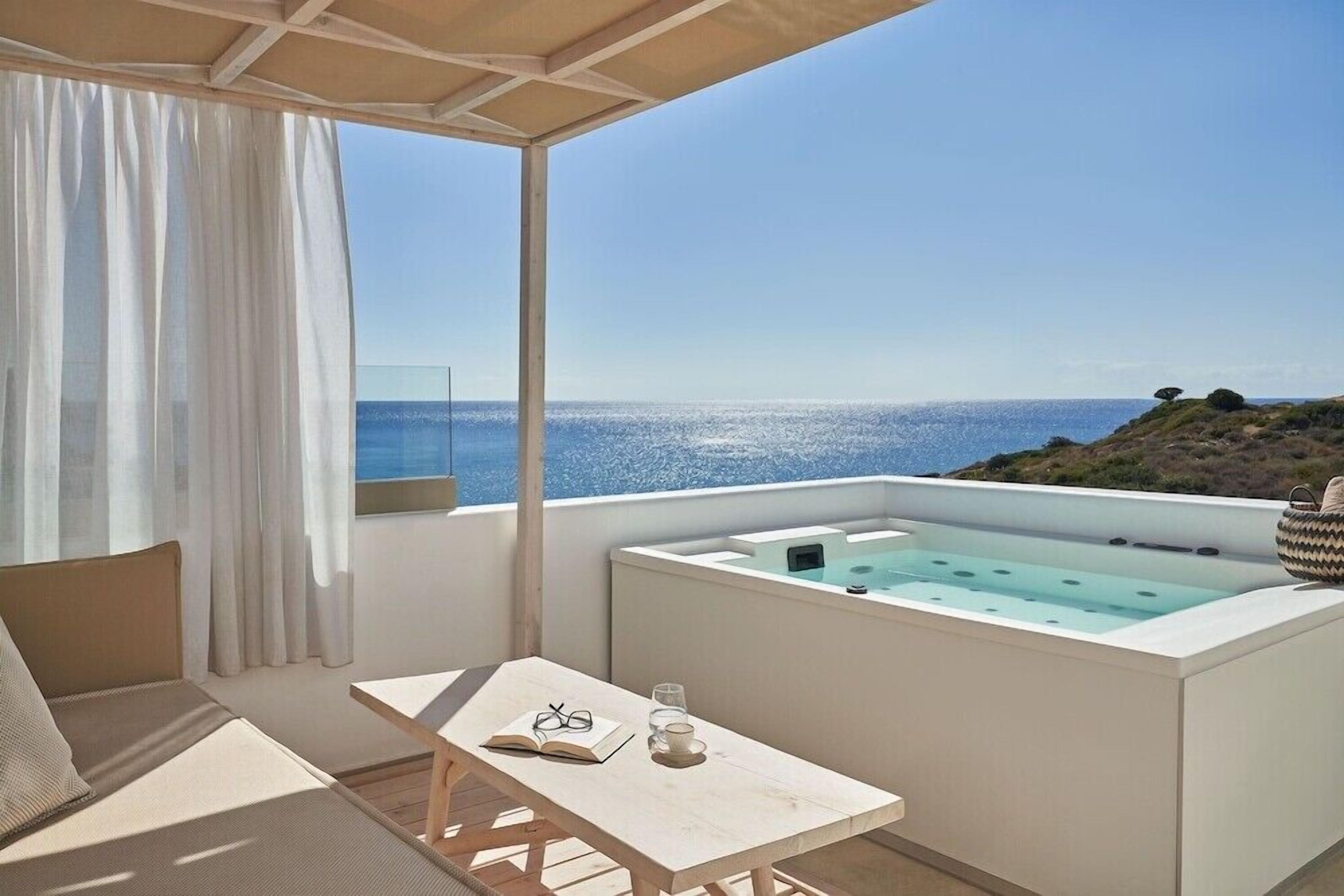 Een balkon met uitzicht op zee, een jacuzzi, een tafel met een boek en glazen, en een bank.