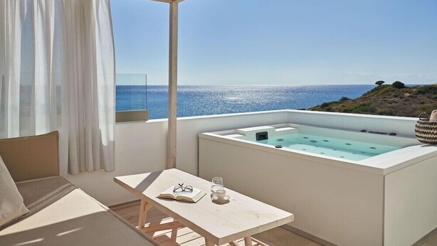 Een balkon met uitzicht op zee, een jacuzzi, een tafel met een boek en glazen, en een bank.