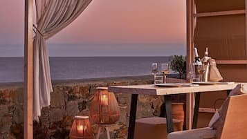 Een terras met uitzicht op zee, een gedekte tafel met glazen en flessen, rieten stoelen en een gordijn.