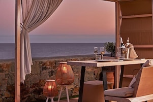 An outdoor dining area with a view of the sea, a table set with glasses and bottles, wicker chairs, and a curtain.