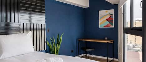 A hotel room with a bed, a desk, a chair, a potted plant, and a window.