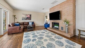 Un salon avec une cheminée en briques, un téléviseur à écran plat fixé au-dessus, un canapé en cuir marron, un fauteuil bleu et un tapis à motifs.