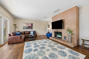 A living room with a brick fireplace, a flat-screen TV mounted above it, a brown leather sofa, a blue armchair, and a patterned rug. - Waterfront Amelia Island home with amazing views, deck, parking. (Fernandina Beach)