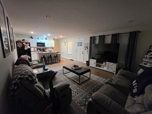 Een woonkamer met een bank, een salontafel en een tv-meubel. Achtergrond is een keuken met een koelkast en een magnetron.