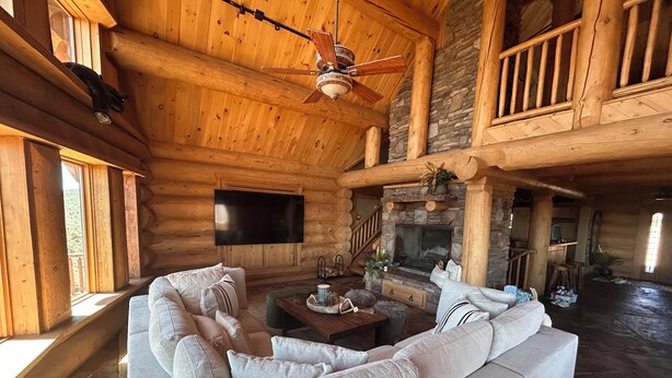 Interior de una cabaña de troncos con sofá seccional, chimenea, televisor y techo de madera.
