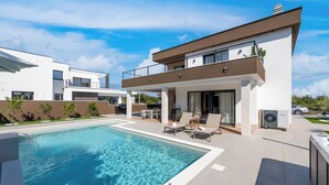 Et moderne hus i to etager med swimmingpool, udendørs møbler og en synlig aircondition.