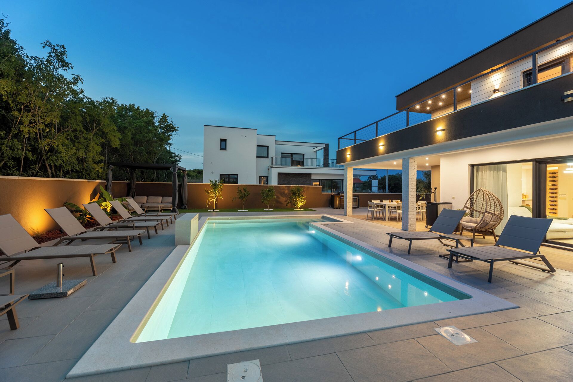 A modern outdoor pool area with lounge chairs, a barbecue, and a two-story building in the background.