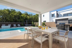 Ett modernt utomhusområde med plats för måltider, en pool, en grill och en överbyggd sittplats.