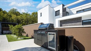 Et udendørs køkken med grill, ovn og højtalere, beliggende ved siden af et moderne hus med en balkon.