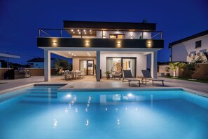 Ett modernt hus i två våningar med en swimmingpool, utomhusmöbler och en väl upplyst uteplats.