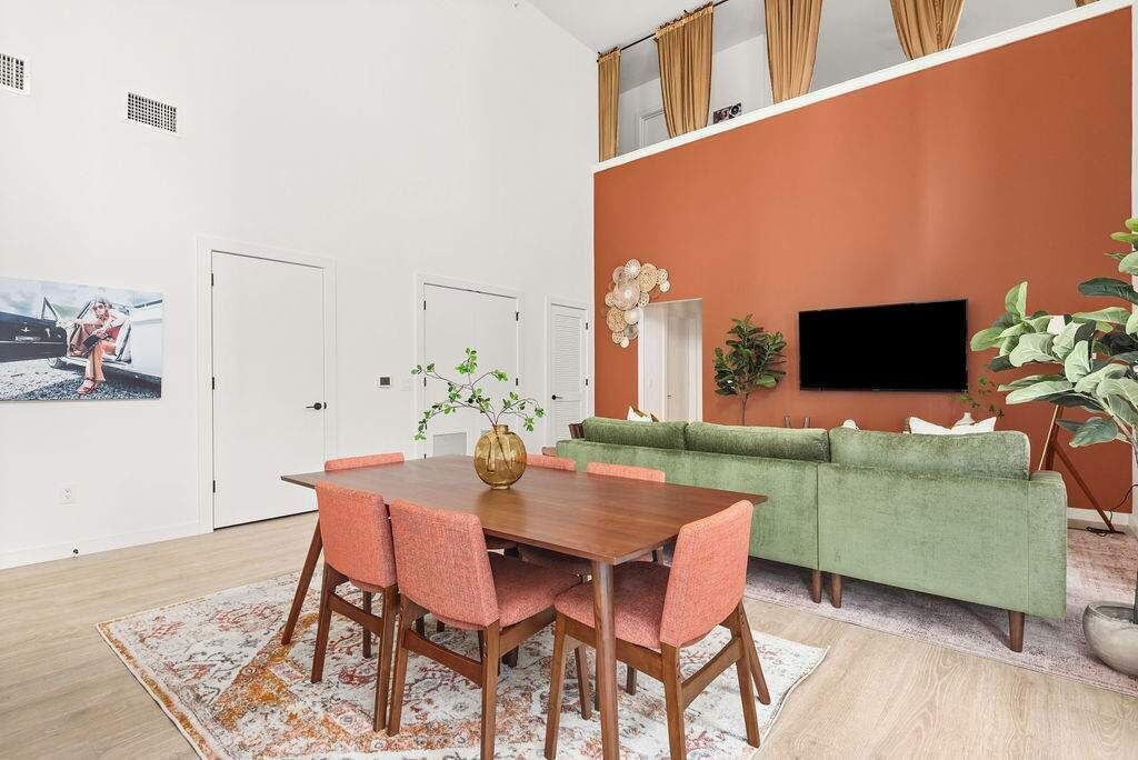 Een eetgedeelte met een houten tafel en roze stoelen, een groene bank en een televisie aan de oranje wand.