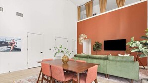 Et spiseområde med et træbord og pink stole, en grøn sofa og et fjernsyn monteret på den orange væg.