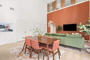 Ein Essbereich mit einem Holztisch und rosa Stühlen, einer grünen Couch und einem an der orangefarbenen Wand befestigten Fernseher.