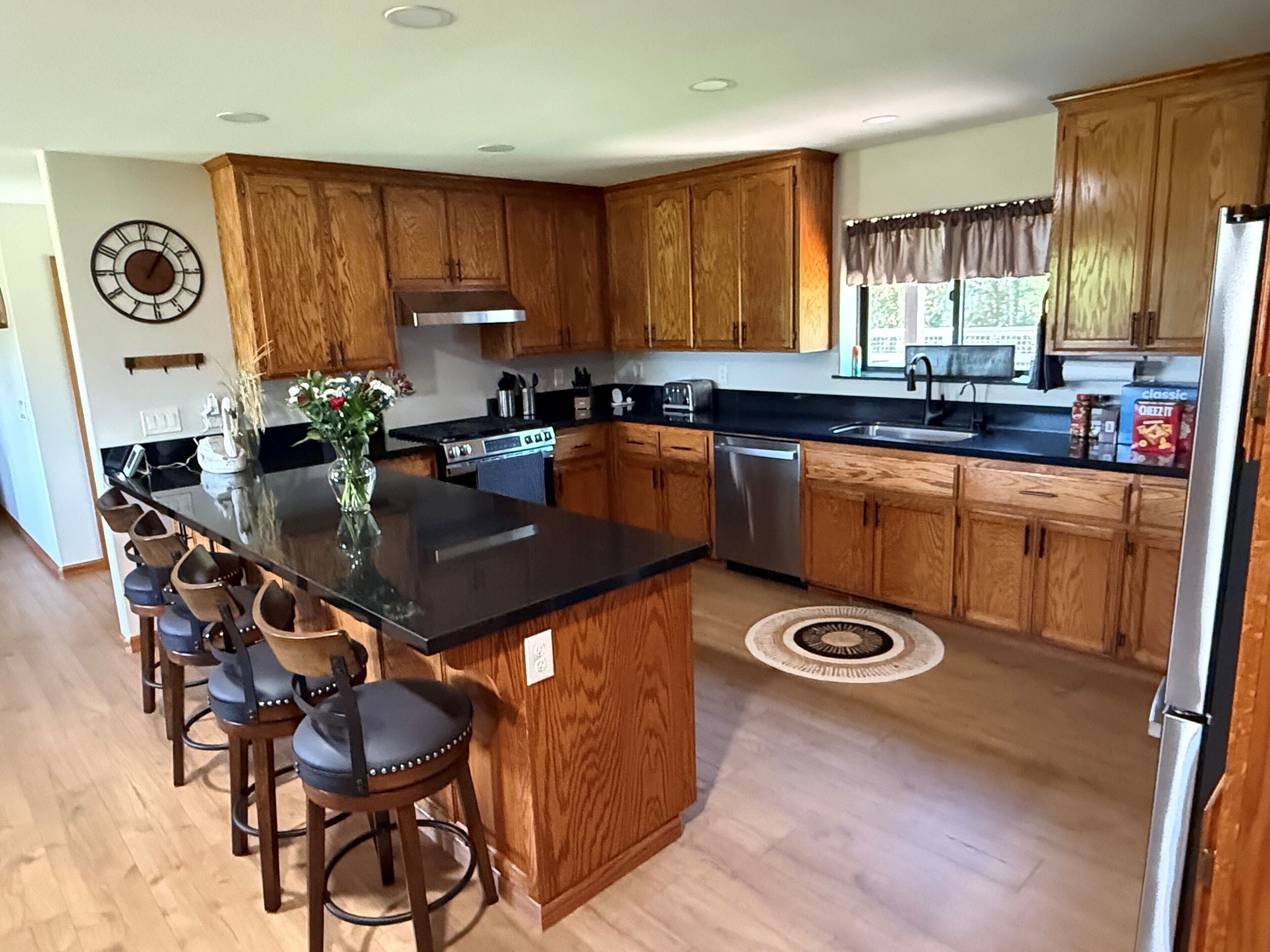 A modern kitchen with wooden cabinets, a central island with bar stools, stainless steel appliances, and a clock on the wall.