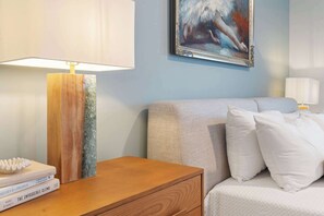 A neatly arranged bedroom with a wooden nightstand, a bedside table, a bed with white bedding, and a painting on the wall. - The Grand Skyline | 2 Designer Condos, Pool, Gym (Nashville)
