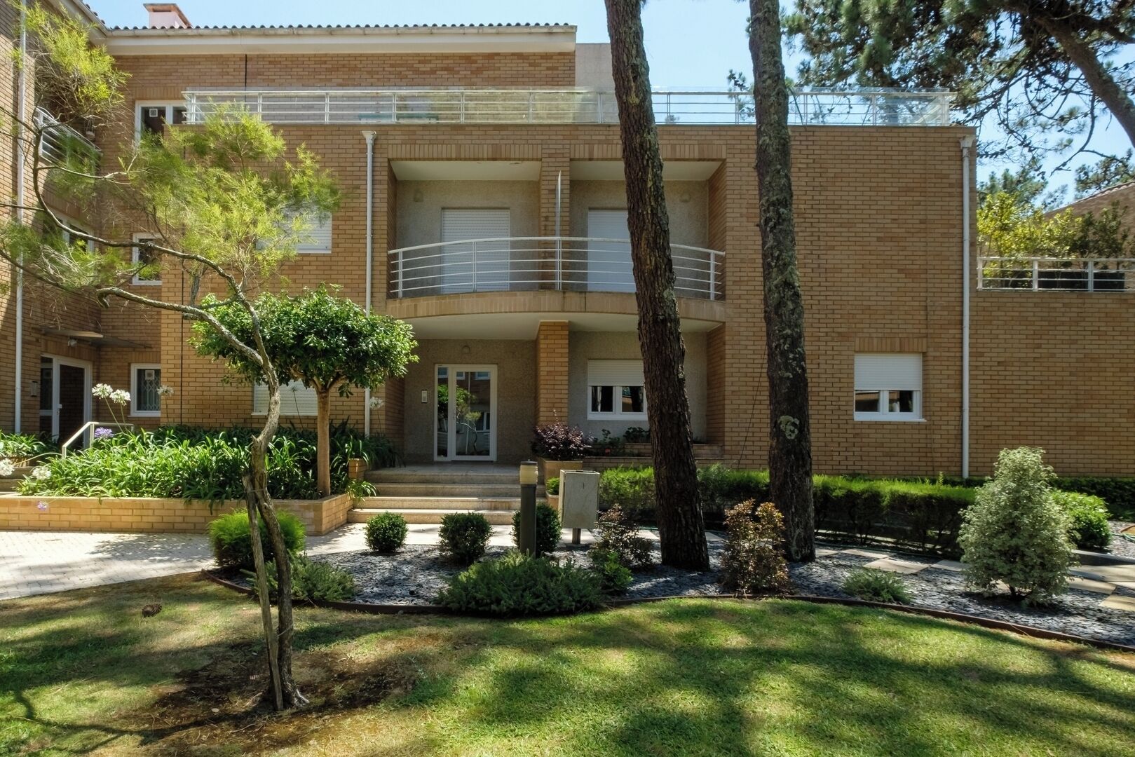 A two-story building with a balcony, a small garden area with trees and shrubs, and a paved entrance.