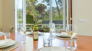 A dining table set for four with wine glasses, plates, and cutlery, a bottle of wine, and a view of trees through large windows.