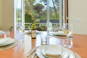 A dining table set for four with wine glasses, plates, and cutlery, a bottle of wine, and a view of trees through large windows.