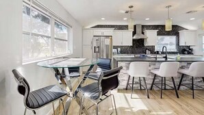 Cocina moderna con armarios blancos, salpicadero negro y electrodomésticos de acero inoxidable. Cuenta con un comedor con mesa de vidrio y sillas metálicas.