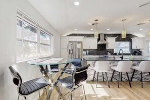 A modern kitchen with white cabinets, a black backsplash, and stainless steel appliances. There is a dining area with a glass table and metal chairs. - Newly Renovated Beach Retreat Just Steps from Englewood Beach - Beach Barracks - Roelens (Englewood)
