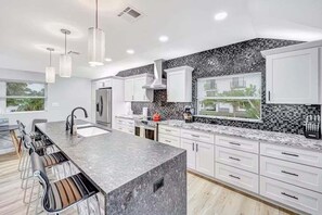 A modern kitchen with a large island, white cabinetry, and a black and white backsplash. - Newly Renovated Beach Retreat Just Steps from Englewood Beach - Beach Barracks - Roelens (Englewood)
