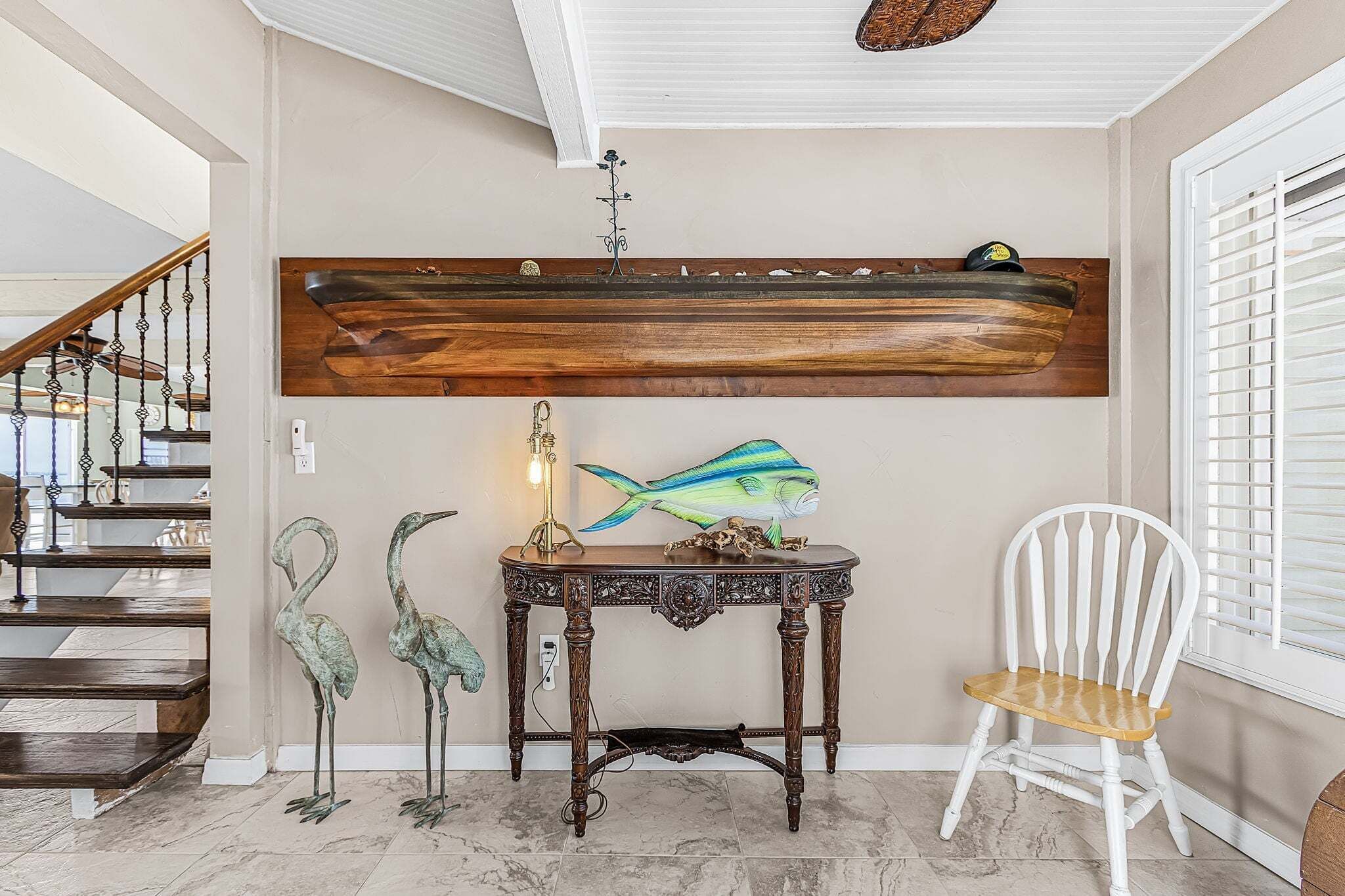 A staircase with a wooden handrail, a wall-mounted shelf with a decorative item, a table with a lamp, and a white chair.