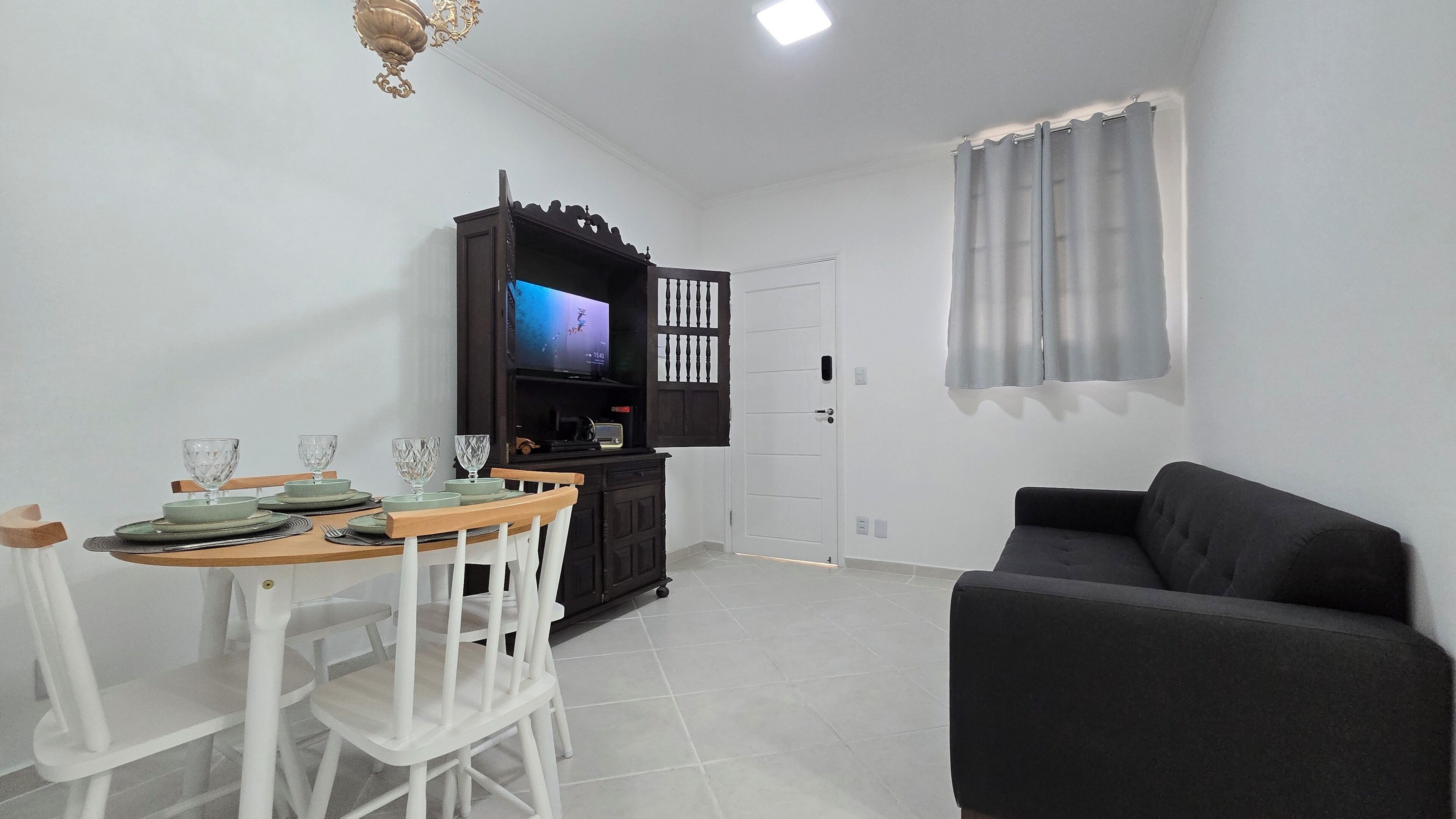 Uma sala de estar com um sofá preto, uma mesa de jantar com cadeiras, uma TV em um armário de madeira e uma porta branca.