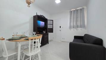 Uma sala de estar com um sofá preto, uma mesa de jantar com cadeiras, uma TV em um armário de madeira e uma porta branca.