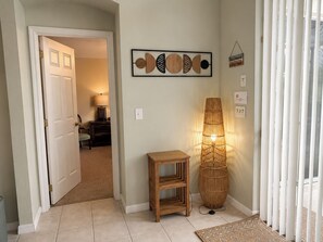 Ein Flur mit einer Tür zu einem Zimmer, einem an der Wand befestigten abstrakten Kunstwerk, einer Stehlampe und einem kleinen Holztisch.