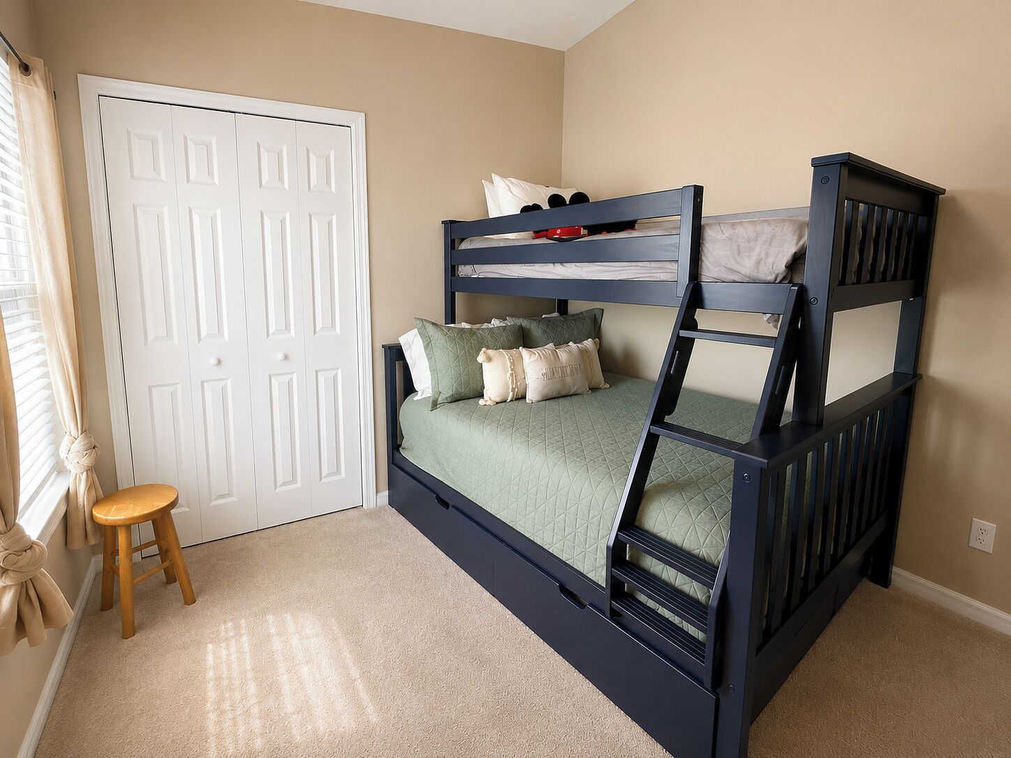 A bunk bed with a wooden ladder, a small stool, and a white door in a room with beige walls.