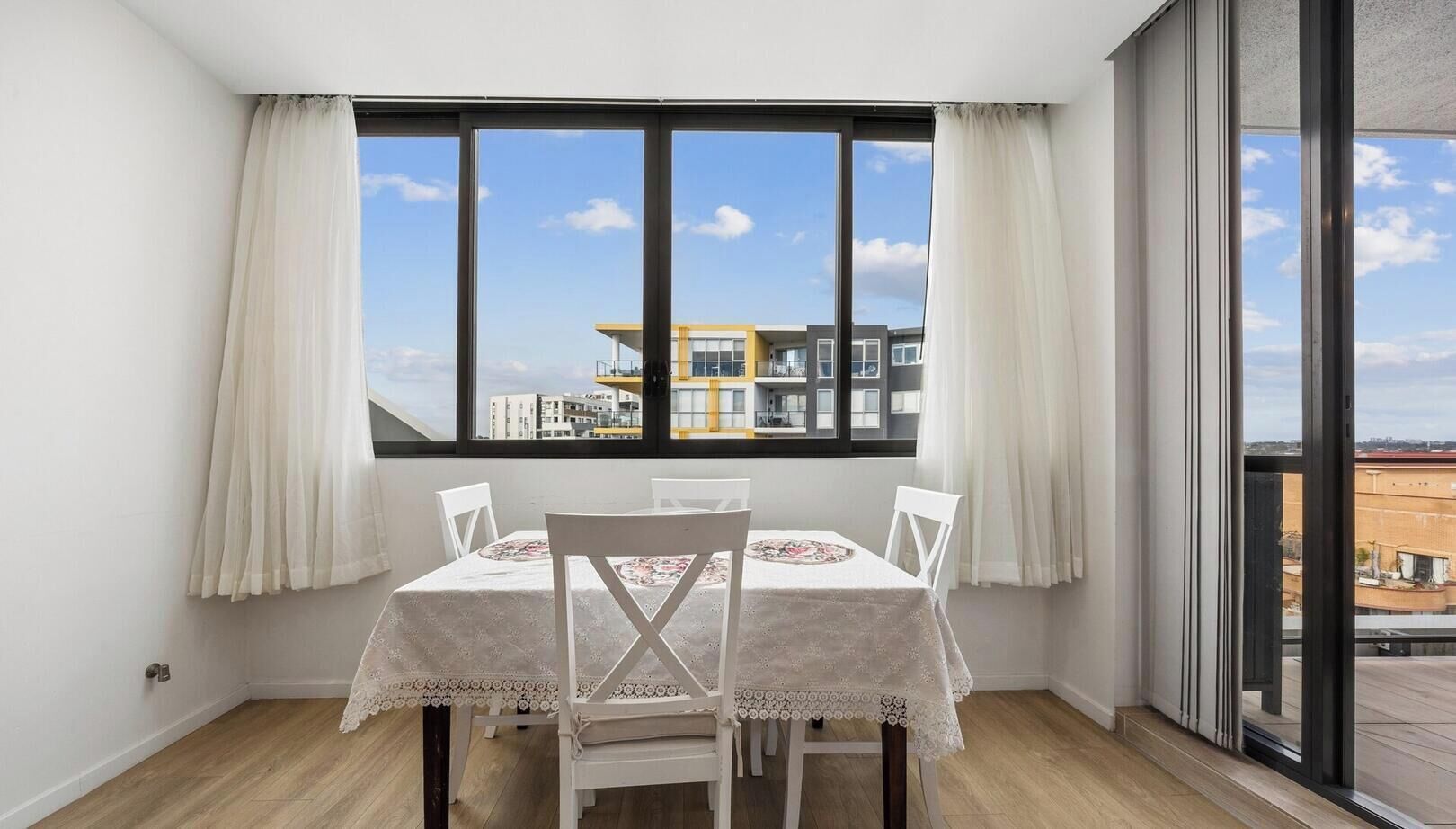 Ein Essbereich mit Tisch für vier Personen, großen Fenstern mit Vorhängen und Blick auf Gebäude.