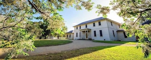 Um edifício grande de dois andares com fachada simétrica, entrada central com colunas e um jardim ao redor.