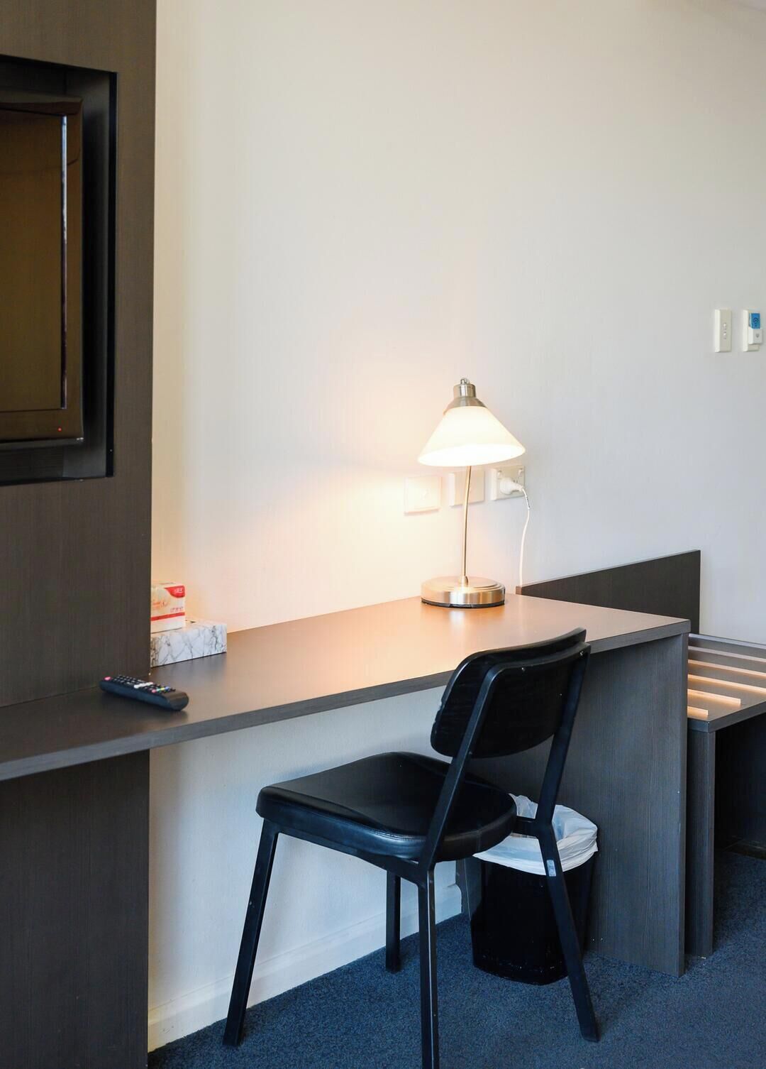 Et hotelværelse med et skrivebord, en stol, en lampe, en fjernbetjening og en skraldespand.