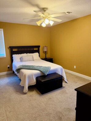 Ein Schlafzimmer mit einem Bett, einem Nachttisch, einer Lampe, einem Deckenventilator und einem Fenster mit Jalousien.