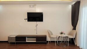 A modern living room with a flat-screen TV mounted on the wall, a wooden entertainment unit, two upholstered chairs, a small round table with a vase, and a window with curtains.