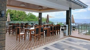 An outdoor dining area with tables and chairs, a view of the sea, and a covered section with a railing.