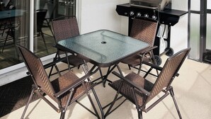 Um pátio externo com mesa de vidro, quatro cadeiras, ventoinha no teto e churrasqueira.