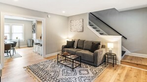 Una sala de estar con un sofá gris, una mesa de centro, un tapete estampado, una escalera y un comedor con sillas y una mesa.