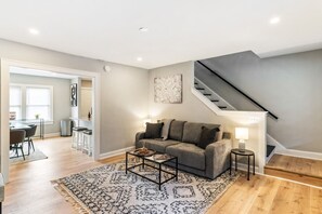 A living room with a grey sofa, a coffee table, a patterned rug, a staircase, and a dining area with chairs and a table. - Pet-Friendly 2BR in Merion Village (Columbus)