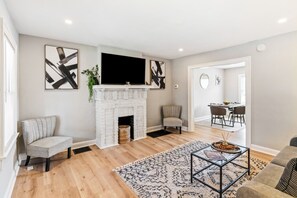 A living room with a fireplace, a TV mounted above it, a dining area in the background, and a sofa. - Pet-Friendly 2BR in Merion Village (Columbus)