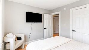 Un dormitorio con cama, una silla, televisor montado en la pared y una puerta que da a otra habitación.