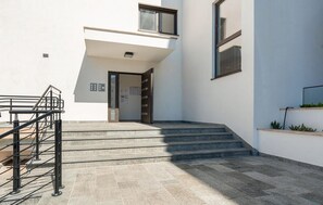 Ein moderner Gebäudeeingang mit einer Treppe, die zu einer Tür führt, einem kleinen Außenbereich mit Stufen und einem Metallgeländer.