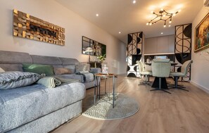 Een moderne woonkamer met een grijze bank, een eethoek met een tafel en stoelen, en een keuken op de achtergrond.