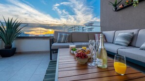 Une terrasse sur le toit avec un canapé, une table à manger garnie de plats et de boissons, et une vue sur le coucher de soleil.