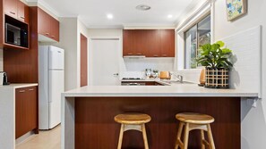 Une cuisine moderne avec des meubles en bois, un four à micro-ondes encastré, un réfrigérateur, une cuisinière et deux tabourets.