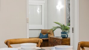 A dining area with a table set for two, a brown leather sofa, a wooden chest of drawers, and a potted fern. - Saltwater Sanctuary | Fireplace & Walk to Beach (Mount Martha)